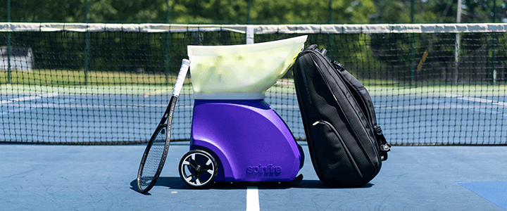 Spinfire Pro 2 on a tennis court with a racquet and tennis bag leaning against it